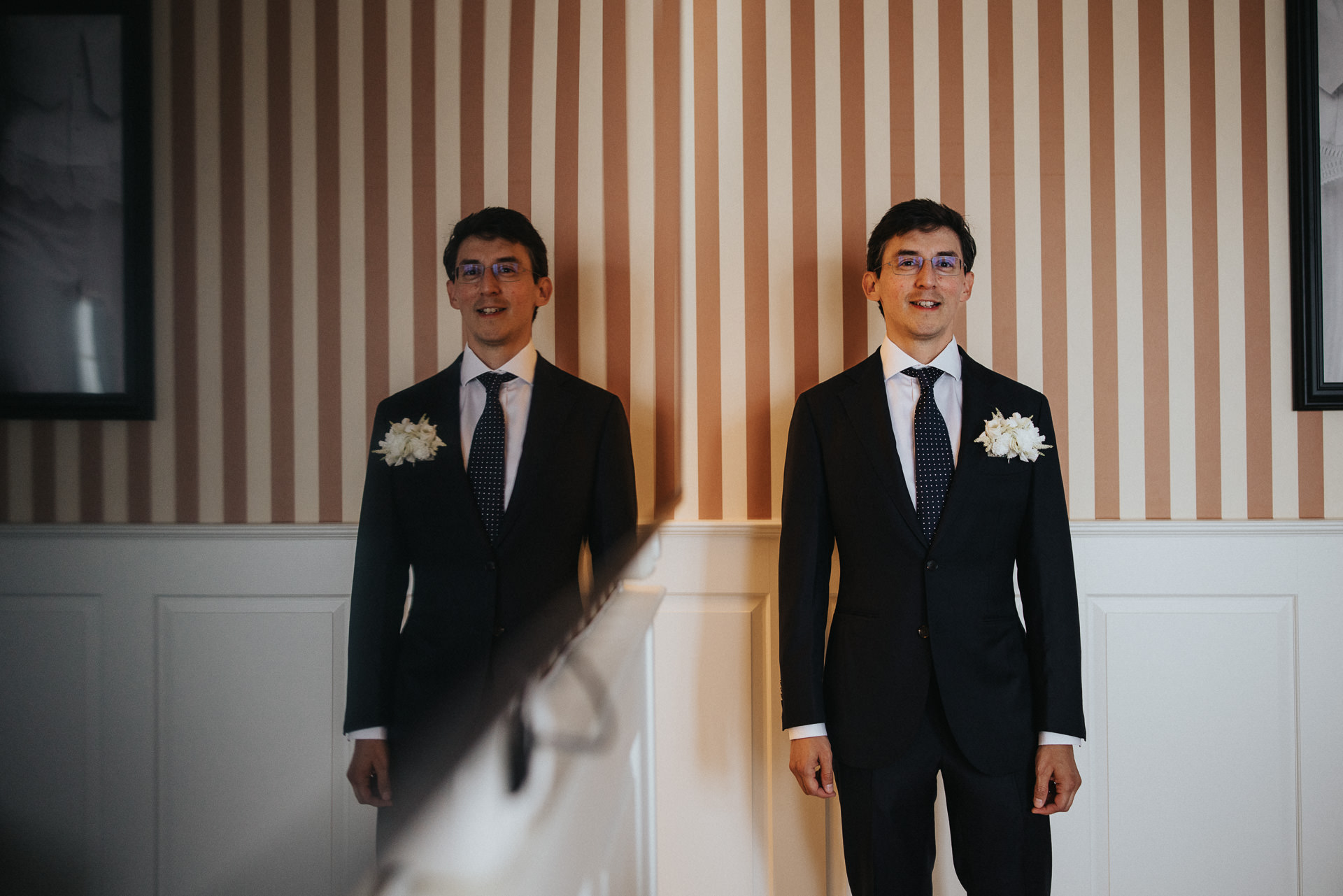 Brudgum i mörk kostym med vit knapphålsblomma framför randig vägg