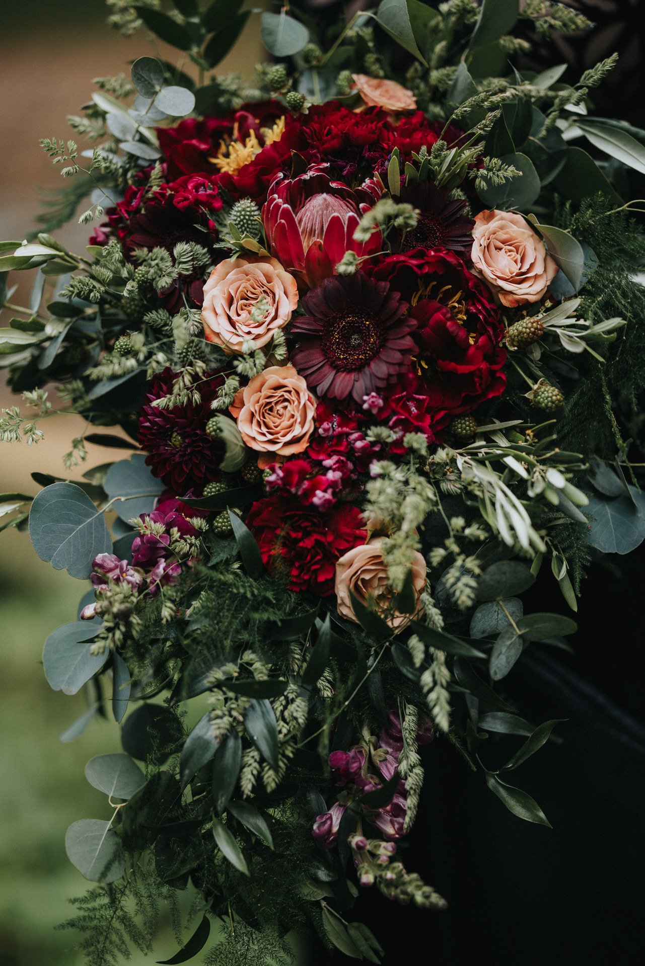 Dramatisk brudbukett med vinröda blommor, aprikosfärgade rosor och gröna blad vid bröllop i Skåne