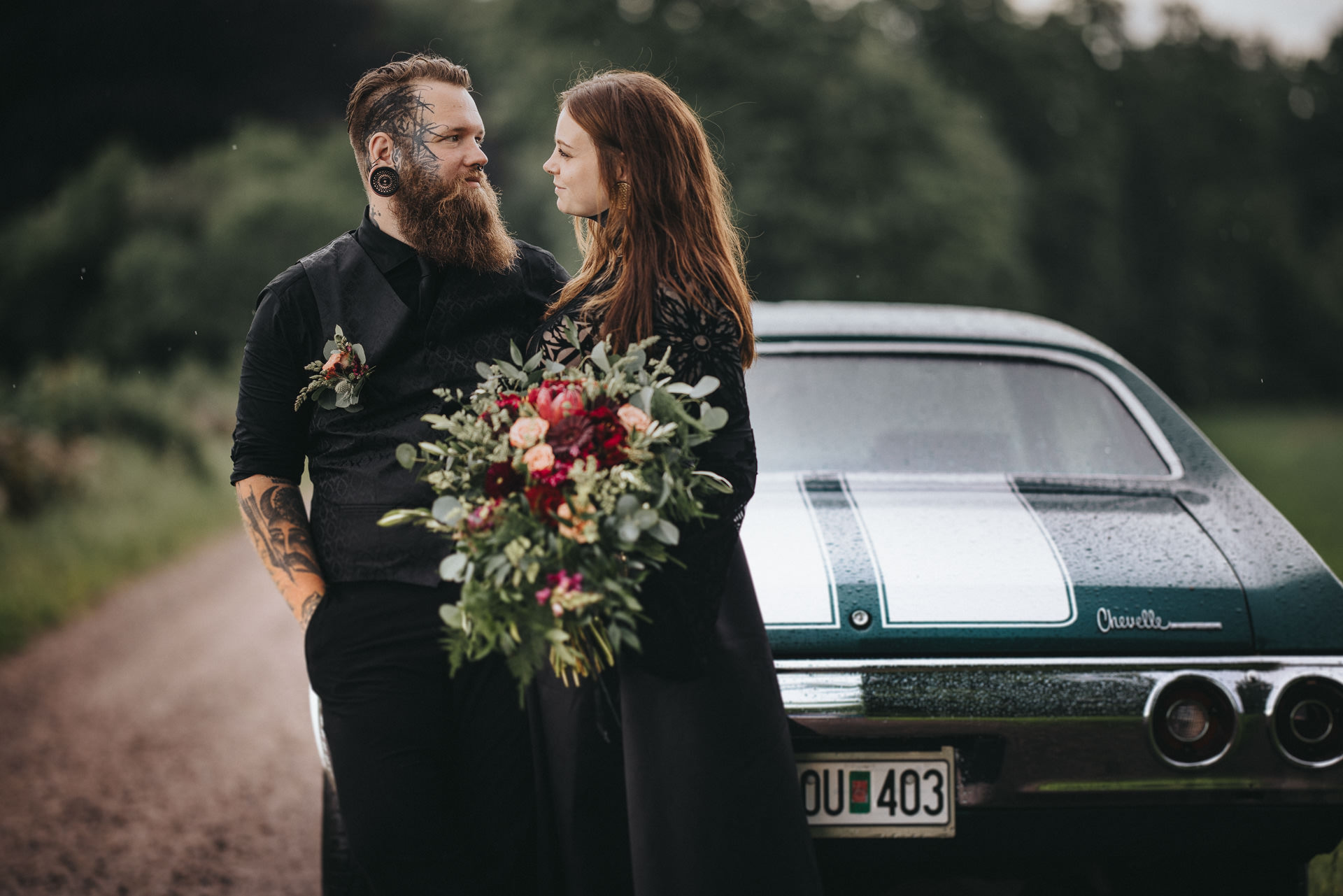 Alternativt brudpar med tatueringar och mörka bröllopskläder framför en klassisk Chevrolet Chevelle i Hässleholm